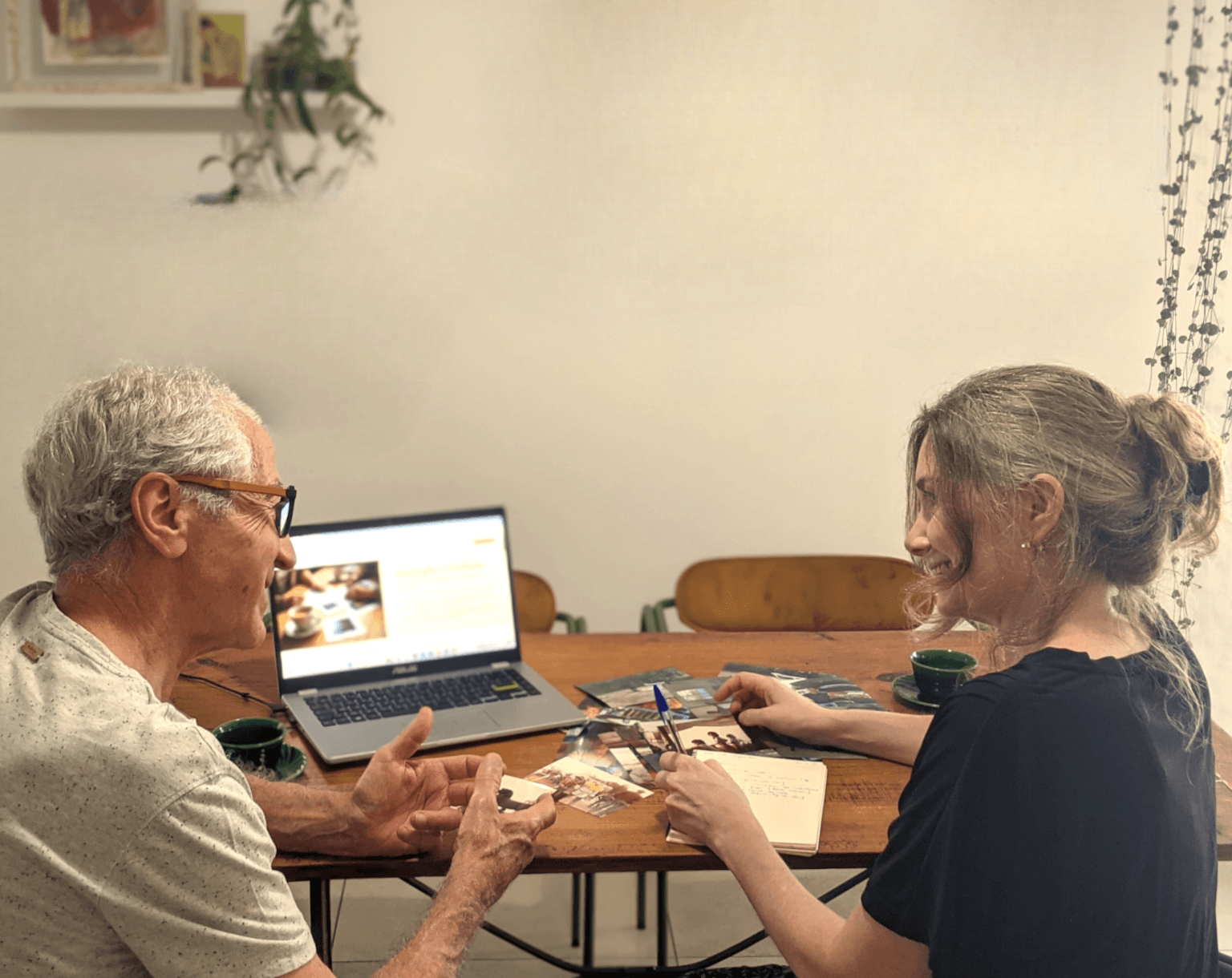 Deux personnes sont assises devant une table. Sur la table il y a des photos et un ordinateur. Les personnes échangent et sourient.