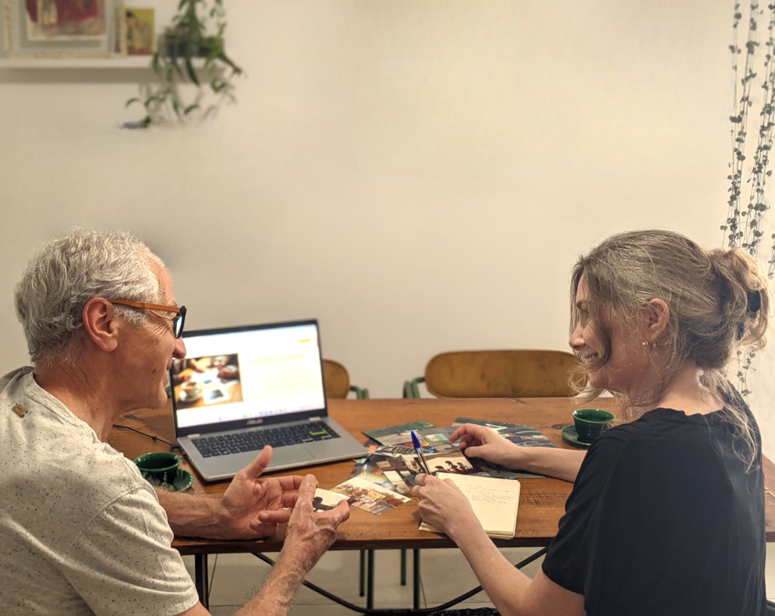 Deux personnes sont assises devant une table. Sur la table il y a des photos et un ordinateurs. Les personnes échangent et sourient.