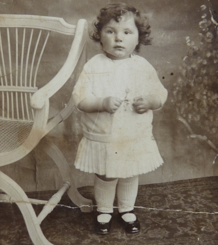 Monique est bébé et se tient debout. Elle porte une robe blanche et des chaussures vernies.