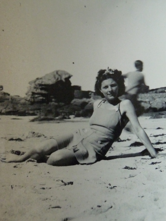 Monique est assise sur la plage en appui sur les bras. Elle porte une robe à bretelles et un nœud dans les cheveux. La photo est en noir et blanc.